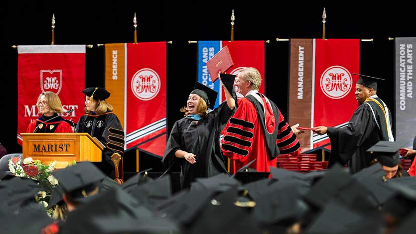 Image of student holding up diploma on stage alongside President Weinman at graduate commencement ceremony.