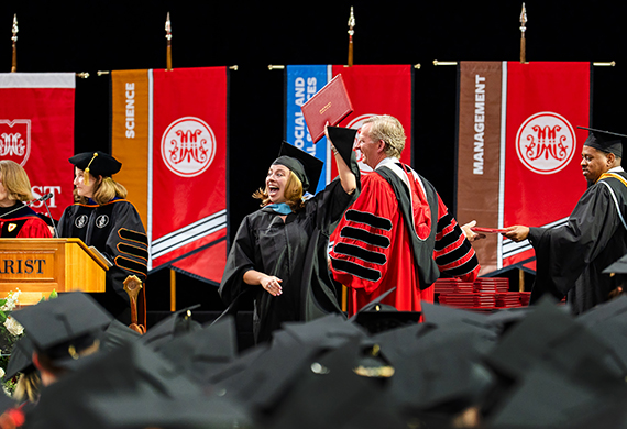 Image of student holding up diploma on stage alongside President Weinman at graduate commencement ceremony.