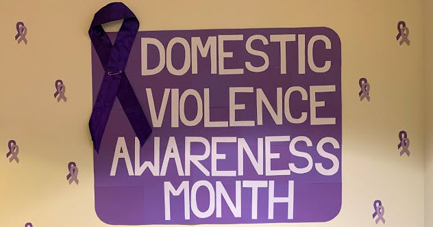  A purple sign with the words Domestic Violence Awareness Month is on a beige wall. There's a large purple ribbon on the left hand side of the poster, plus smaller purple ribbons around the sign on the wall. 