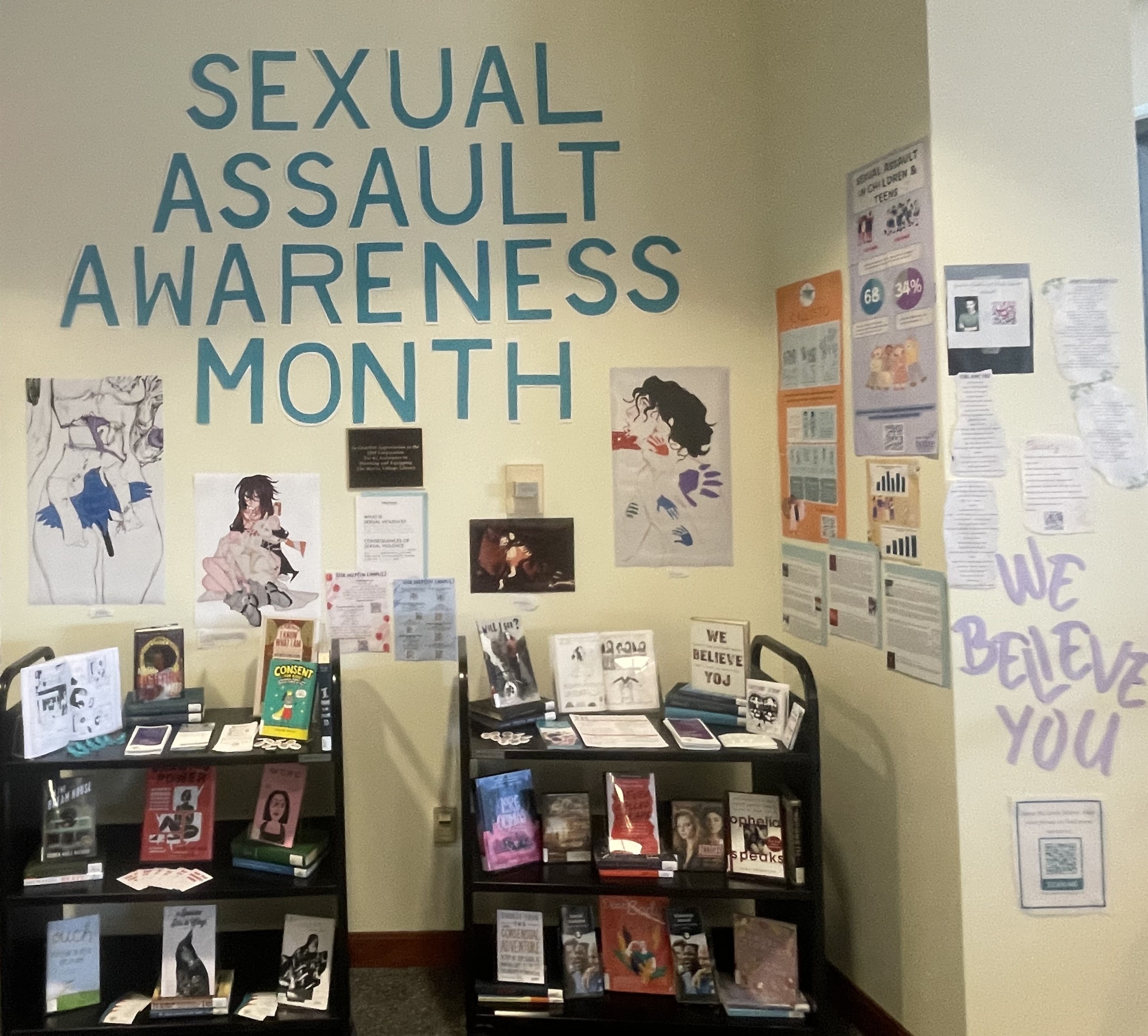 A display of books on carts with the words Sexual Assault Awareness Month on the wall behind the carts. Features artwork, infographics, and poetry underneath the letters. We Believe You is on the right underneath the poetry