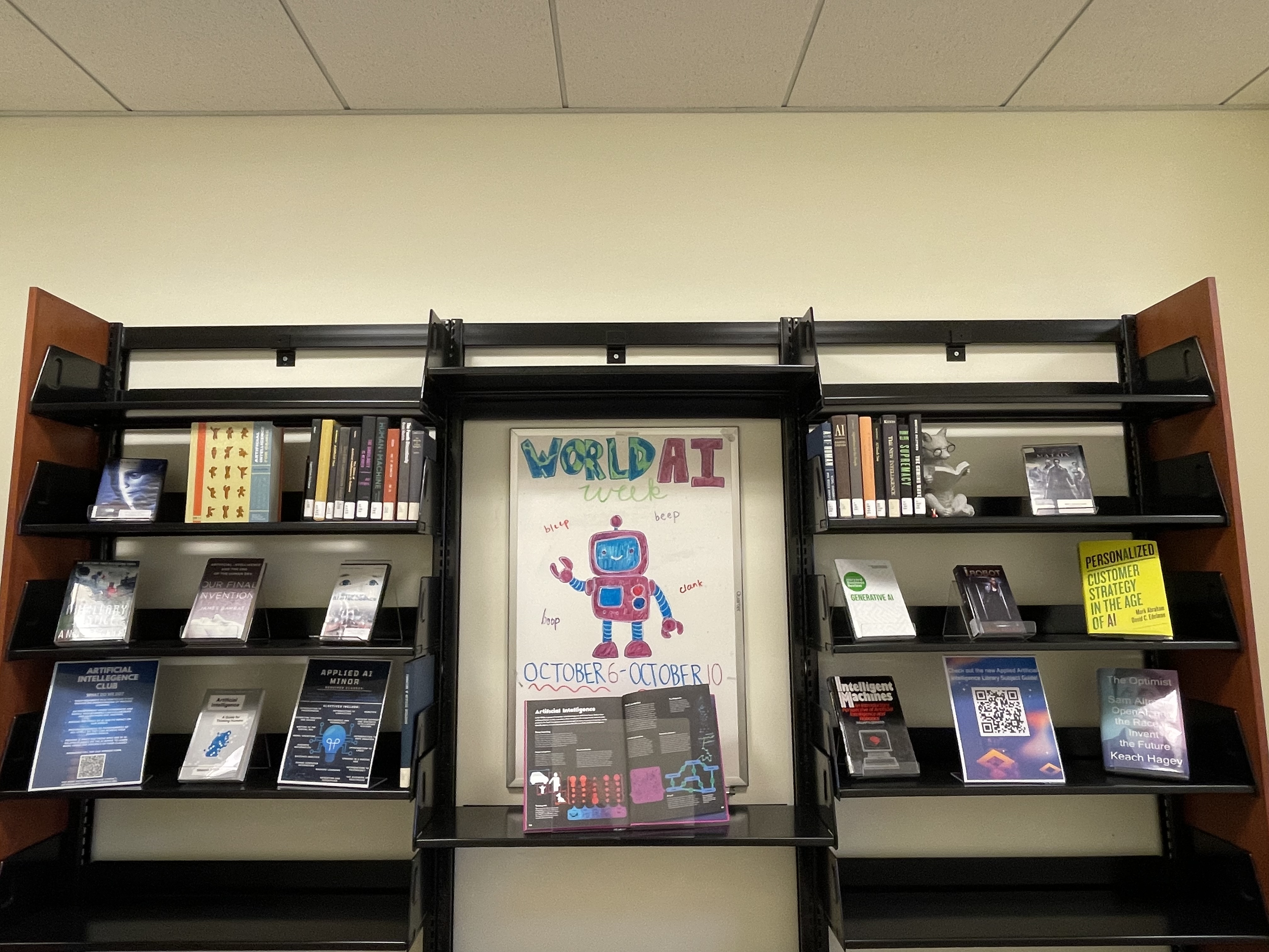  Library shelves with fiction books, nonfiction books, and DVDs that focus on AI. In the center of the shelves is a whiteboard that says "World AI Week: October 6-October 10". There is a robot drawn on the whiteboard. 